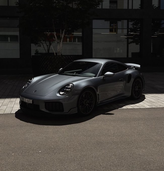 A gray Porsche 911 Turbo (or similar 911 model) sports car parked on pavement in front of a modern building with large windows.