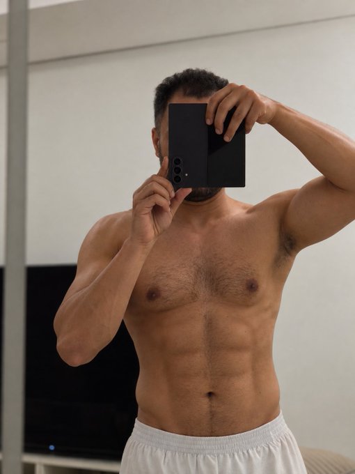 A shirtless man takes a mirror selfie with a black smartphone, holding it up to cover his face while showcasing his toned physique and six-pack abs. The post's caption "Haters will say its AI" humorously plays on the natural skepticism toward unrealistically fit body photos, hinting that the impressive muscular definition might be enhanced or digitally altered.