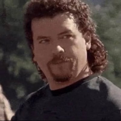 A man with curly brown hair, a mustache, and a goatee is shown in an outdoor setting, looking off to the side.