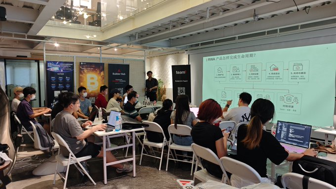 Attendees at Team1 Builder Day in Shenzhen gather in a collaborative workspace, watching a presentation slide titled "RWA 产品怎样完成生命周期的？" (How does an RWA product complete its lifecycle?) displayed on a large screen, while participants work on laptops and engage with the session. The event, branded with "Builder Hub" and "team1" signage, reflects a focused tech and crypto-focused builder gathering.