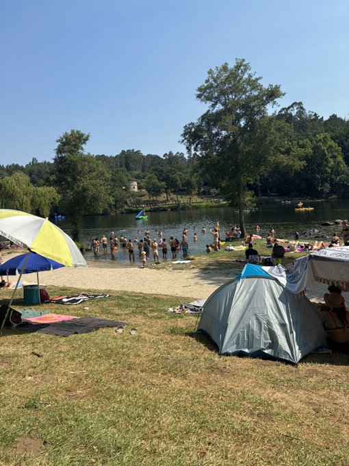 A crowded beach scene at a lake where people are swimming and relaxing under sunny skies, with colorful beach umbrellas and a tent set up on the grassy shore.