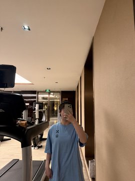 A mirror selfie of a person wearing a light blue Under Armour t-shirt standing in what appears to be a gym or lobby area.