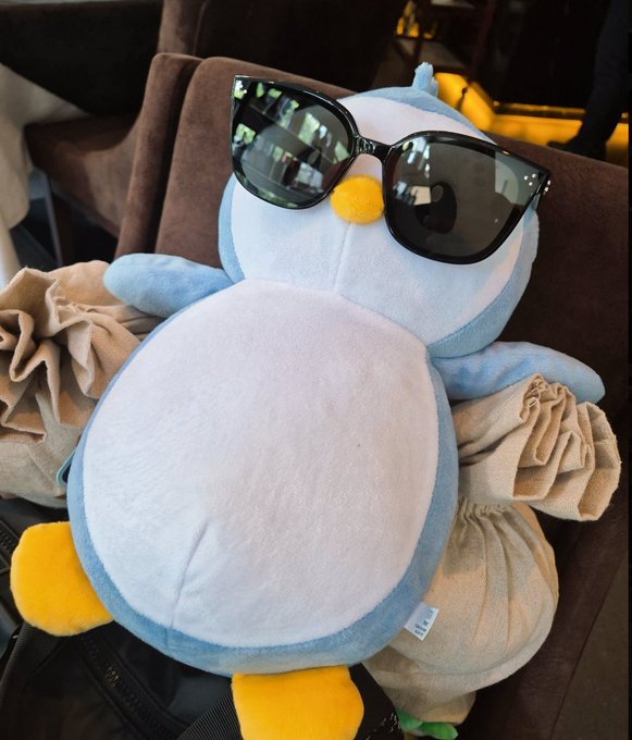 A blue and white plush bird toy wearing large black sunglasses rests on brown cushions, appearing to lounge.