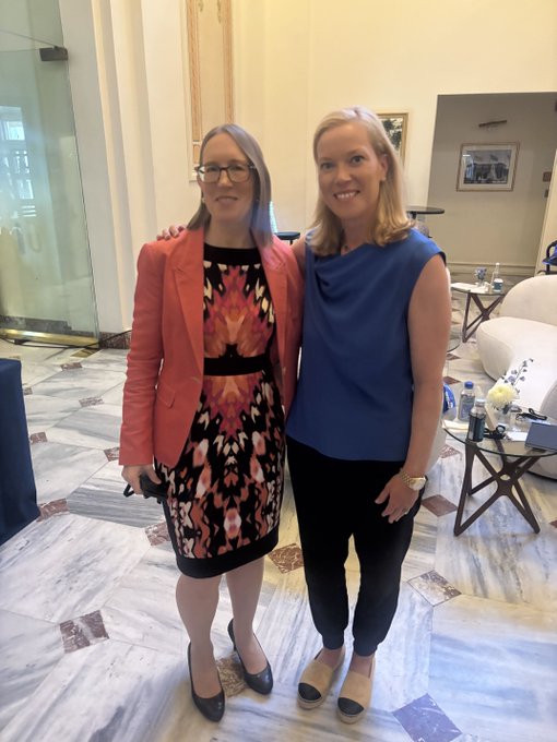 A photograph of two women posing together indoors on a marble floor; the woman on the left wears a floral dress and a coral blazer with glasses, while the blonde woman on the right wears a blue sleeveless top and black pants.