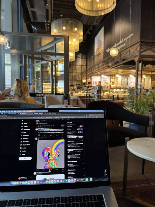 A first-person view of someone working on a laptop at a stylish café, with the screen showing social media feeds including a colorful cartoon character image, while elegant pendant lights and a bar area are visible in the background.