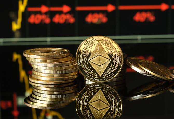 Stack of gold-colored Ethereum Classic coins displayed in front of a blurred red digital chart showing percentage values.