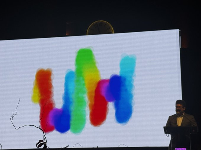 A man stands at a podium in a dark auditorium presenting in front of a massive screen displaying colorful, blurred abstract shapes in red, yellow, green, and blue.