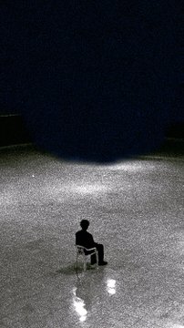 A solitary figure sits on a chair on a vast, illuminated platform in an otherwise dark space, their reflection visible on the glossy floor.