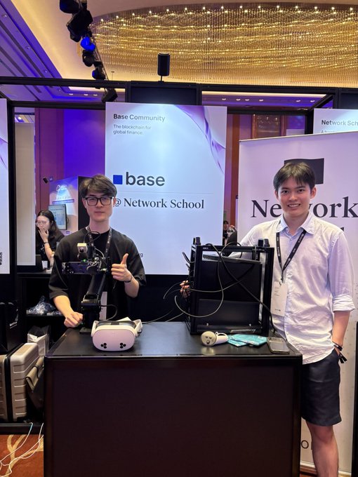 The image features two young men standing next to a booth at a convention or event. The booth displays a large screen with the words "Base" written on it, along with an image of a person holding a microphone. One man is wearing glasses while the other has a lanyard around his neck. They are both smiling and appear to be engaged in conversation or demonstration related to the base's projects.

The background shows several people walking by, indicating that this event is well-attended. The image also contains text about the base's projects on AI and onchain economies, suggesting a focus on technological advancements and innovation within these fields.