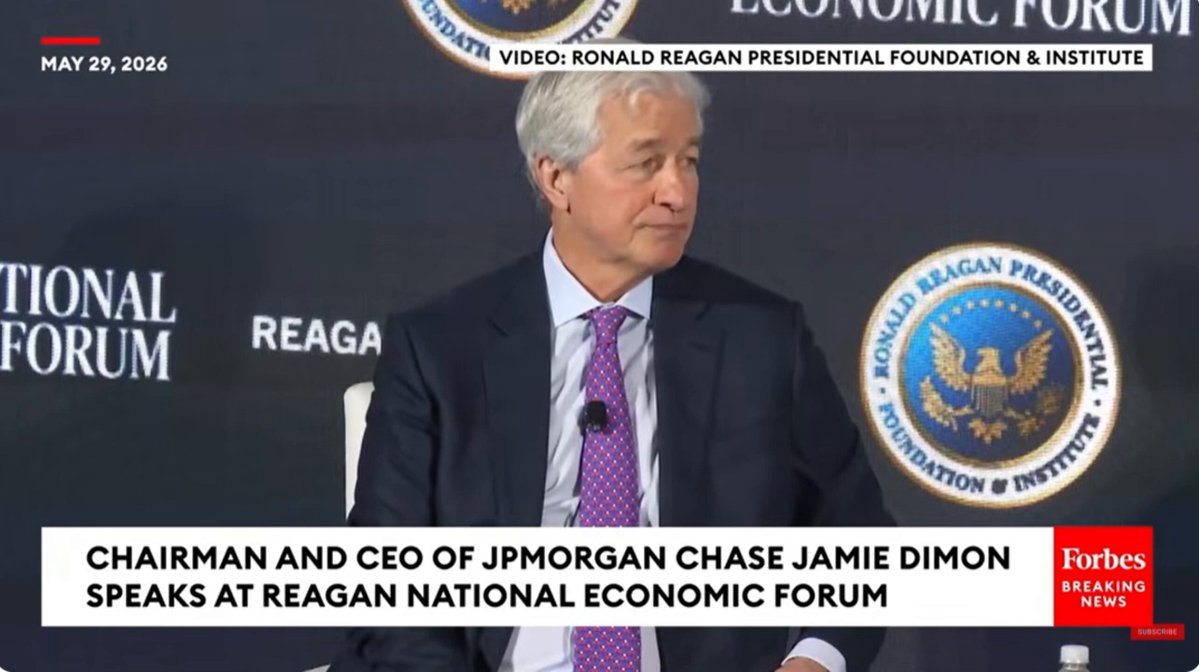 The image shows a man sitting at a table with a podium behind him, likely during a press conference or event. He is wearing a suit and tie, indicating that he holds a high position within an organization. The text "Chairman and CEO of JPMorgan Chase Jamie Dimon speaks at Regan National Economic Forum" suggests that the image might be related to a panel discussion or interview featuring the man in question.