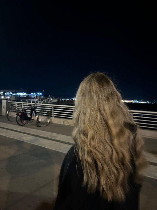 A woman with long, wavy blonde hair stands with her back to the camera, gazing out at city lights across a waterfront at night, with a bicycle parked against the railing nearby. The serene, forward-looking scene echoes the post's optimistic outlook for tomorrow, capturing a quiet moment of nighttime reflection that pairs with the bullish sentiment and "goodnight" sign-off.