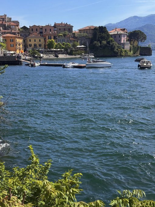 A scenic view of a lake or harbor with boats docked near colorful buildings along the shore, with mountains in the background under a clear blue sky.