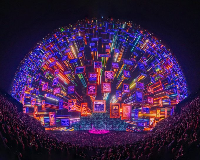 The image shows a large crowd of people gathered at an event, possibly a concert or performance, with many colorful lights illuminating the area. The audience appears engaged and focused on the spectacle before them. The photo has been edited to emphasize the vibrant atmosphere created by the lights and the excitement of the event.