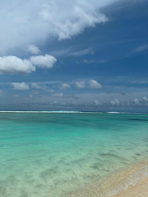 Crystal clear turquoise waters meet a sandy shoreline under a bright blue sky scattered with soft clouds, evoking a perfect "good morning" tropical paradise. The serene beach scene mirrors the uplifting "Gm everyone" greeting with its fresh, sunny start to the day.