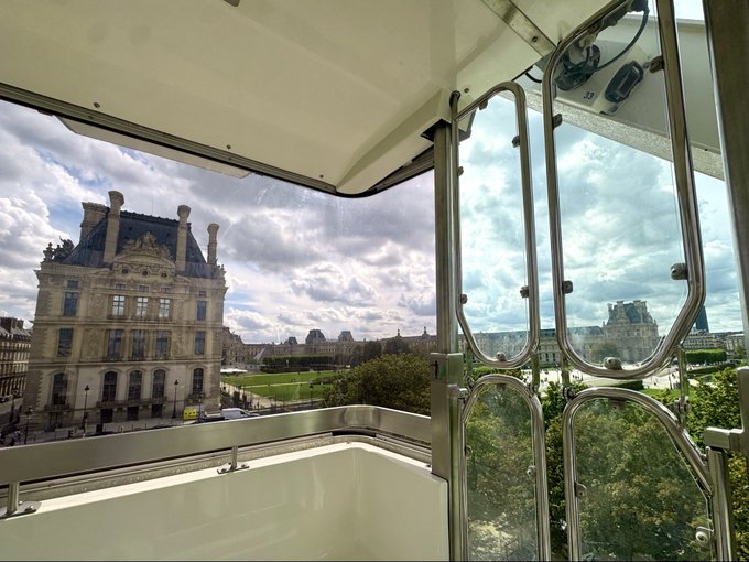 The image shows a view from inside a glass-enclosed cabin or tram looking out toward the Louvre Palace in Paris, with ornate Haussmann-style architecture visible through the windows. The accompanying caption "Checking coins from here" suggests the photo was taken from a vantage point related to the iconic Paris Ferris wheel, likely referring to viewing the city from the Roue de Paris (Grande Roue) ride, though the architectural view prominently features the Louvre's classical façade.