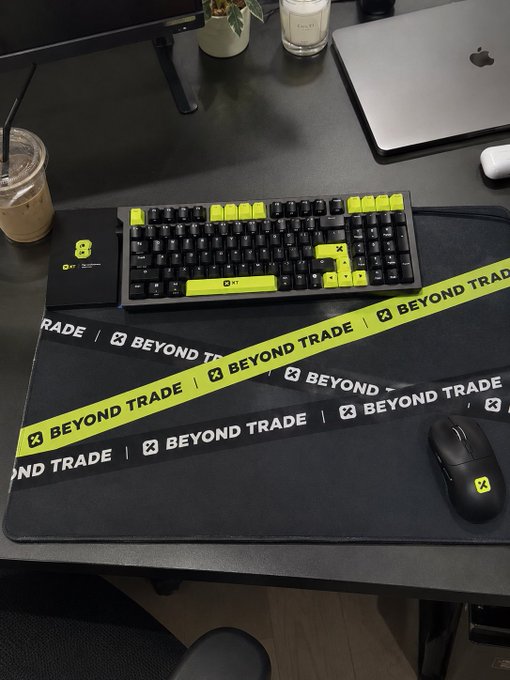 A sleek gaming desk setup featuring a black and yellow "Beyond Trade" branded mousepad, a mechanical keyboard with striking yellow accent keys, a mouse, and a laptop, alongside an iced coffee and plant. The branded gear ties into the post celebrating a $1000 keyboard from @XTexchange and @XTexchangeCN, while wishing followers a Happy Mid-Autumn Festival.