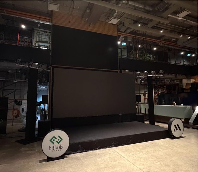 Photo of an empty event stage set up inside an industrial venue, featuring large LED screens, lighting rigs, and a 'bitkub exchange' branded sign at the front of the stage.
