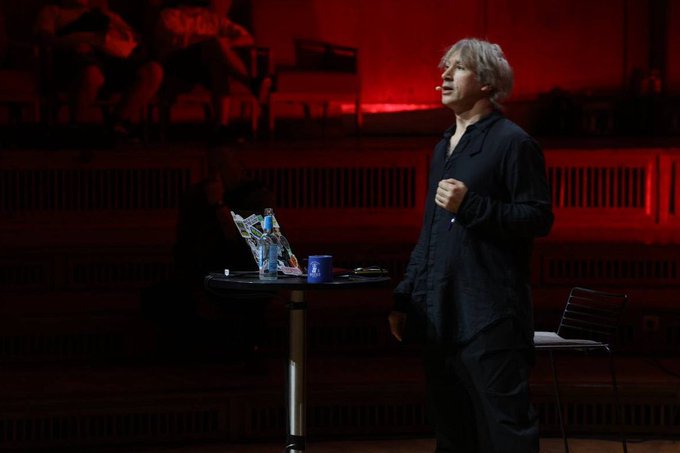 The image shows a man standing at a table with a laptop computer, possibly giving a presentation or lecture. The background features a red wall that contrasts with the gray floor of the room. There are two chairs visible in the scene - one near the center of the room and another closer to the foreground. A cup is placed on the table next to the laptop.