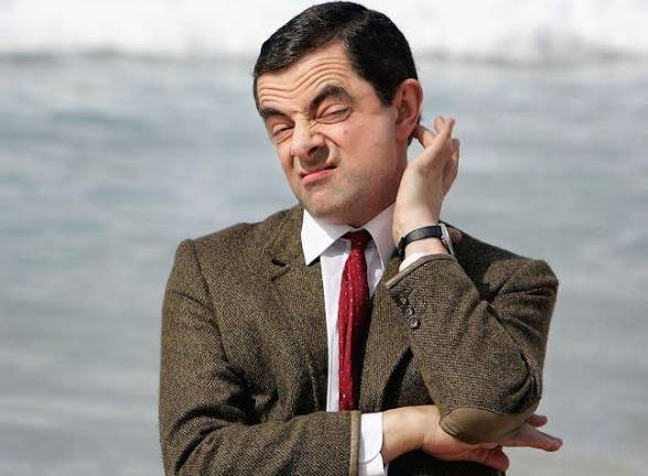 Mr. Bean (Rowan Atkinson) makes a confused, squinting expression while touching his ear, standing outdoors in front of a body of water.