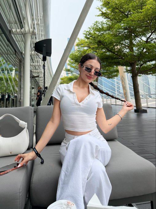 A young woman with long braided hair wearing sunglasses, a white short-sleeved polo crop top, and white pants sits on an outdoor gray sofa, holding her braid, with a white handbag beside her.