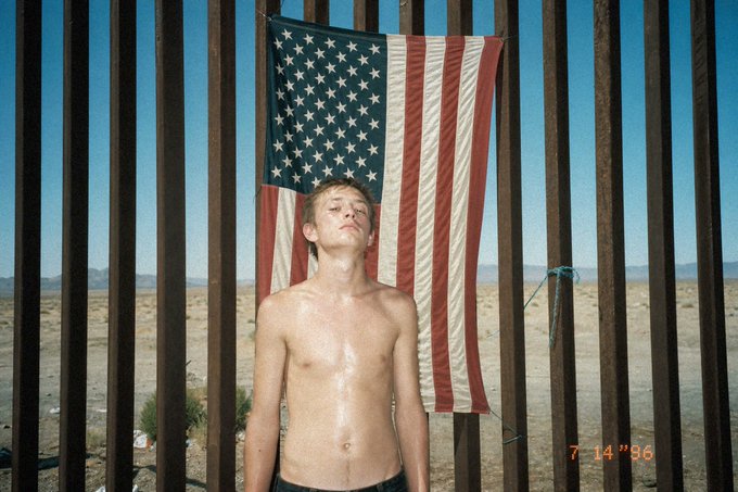 Photograph dated 7/14/96 showing a shirtless young man standing in front of a US flag hung on a metal border fence in a desert landscape.