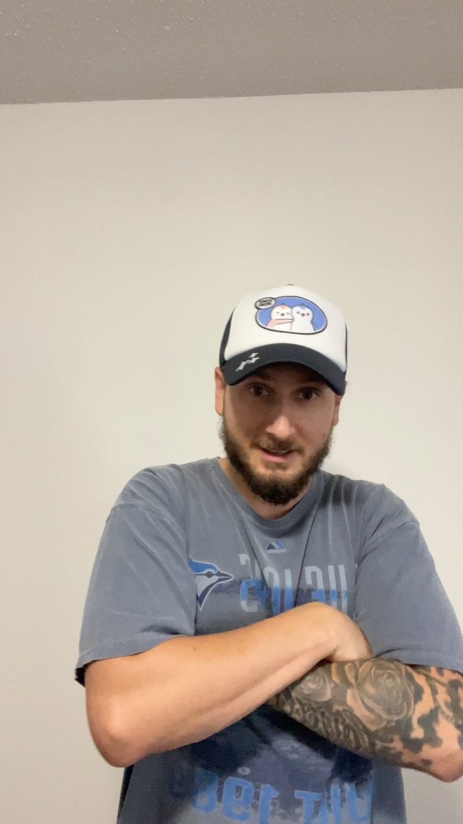 A man with a beard wearing a baseball cap with a cartoon patch and a gray 'Toronto Blue Jays' t-shirt stands with arms crossed, showing a rose tattoo on his forearm.