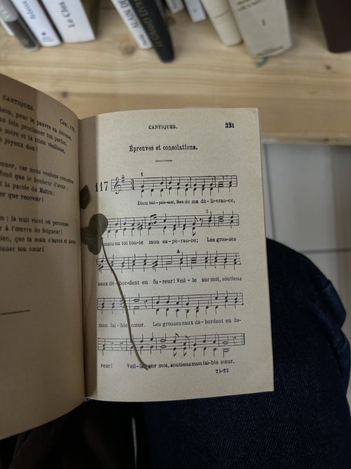 An old French music book lies open on someone's lap, displaying page 331 titled "Épreuves et consolations" with numbered musical notation beneath, while the blurred background reveals a shelf of similar books. The caption "dump saisonnier" (seasonal dump) humorously captures the melancholy tone of the piece's title, suggesting a moment of musical reflection during a quiet, possibly rainy afternoon.