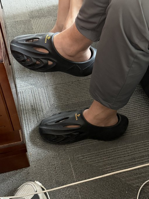 Two black slide sandals featuring the yellow "BINANCE" logo are worn by a person seated on a gray carpet, with one foot propped up. The image showcases the official Binance-branded footwear referenced in the post as crypto-merch seen at a friend's office.