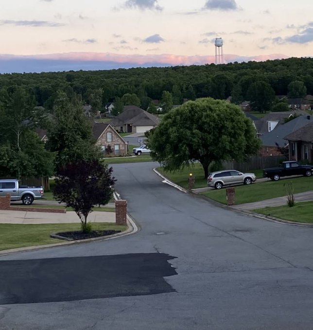 A scenic view of a quiet suburban neighborhood at sunset with houses, trees, parked cars, and a water tower visible in the distance under a pink and cloudy sky.