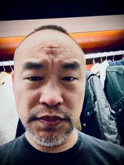 A close-up selfie of a man with short hair and a goatee, with clothing visible on hangers in the background.