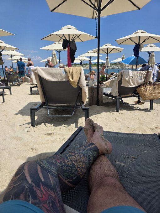 A first-person view from a beach lounge chair, showing tattooed legs in the foreground and rows of white umbrellas and sunbathers on a sandy beach.