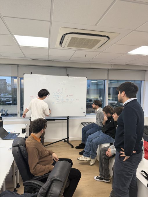 A group of people in an office setting gathered around a whiteboard during a brainstorming or planning session, with one person writing while others observe.