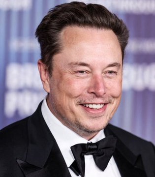 A close-up photo of Elon Musk smiling, wearing a black tuxedo with a bow tie at what appears to be a formal event or gala.