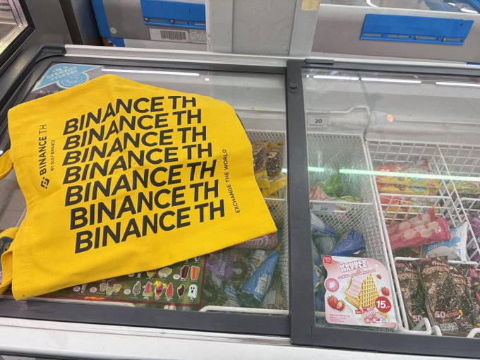 A bright yellow T-shirt with repeated black text reading 'BINANCE TH' and 'EXCHANGE THE WORLD' lies on top of a glass-top freezer filled with various packaged frozen treats.