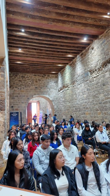A large group of students sits in rows inside a historic stone-walled room with a wooden-beamed ceiling, attending what appears to be a lecture or presentation.