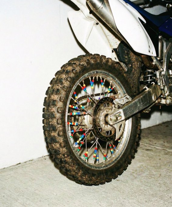 Close-up of a dirty motorcycle's rear wheel with knobby off-road tire, featuring colorful spoke beads in red, blue, yellow, and green.