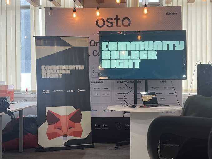 A photo of the MetaMask Community Builder Night event booth, featuring an "osto" branded backdrop and a large TV screen displaying "COMMUNITY BUILDER NIGHT" alongside a retractable banner showcasing partner logos (MetaMask, Linea, OSEN) and the iconic MetaMask fox logo. The image captures the event setup, aligning with the post's mention of a weekend session focused on MetaMask, Web3, and related sub-niches.