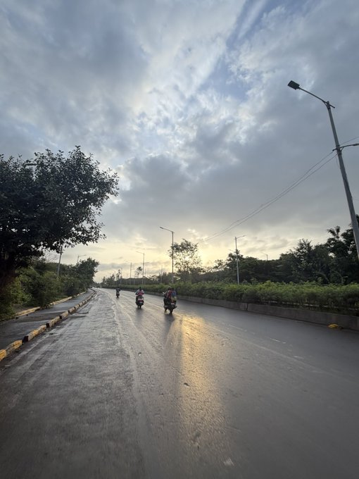 A scenic view of a wet road under a dramatic cloudy sky at sunset, with several motorcyclists riding in the distance.