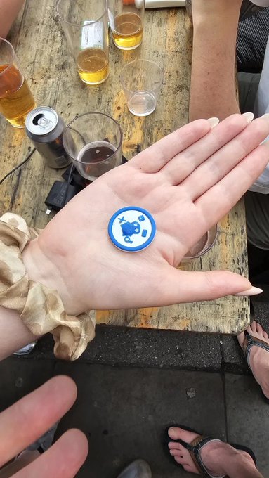 A hand holds up a small, 3D-printed blue and white circular token featuring a cartoon chimp design, displayed against a backdrop of beer glasses and a wooden pub table. The custom NFC collectible chip ties directly to the post's mention of a 3D-printed NFC token created for the Chimpers x Fugz Manchester meetup, where attendees could scan to collect.