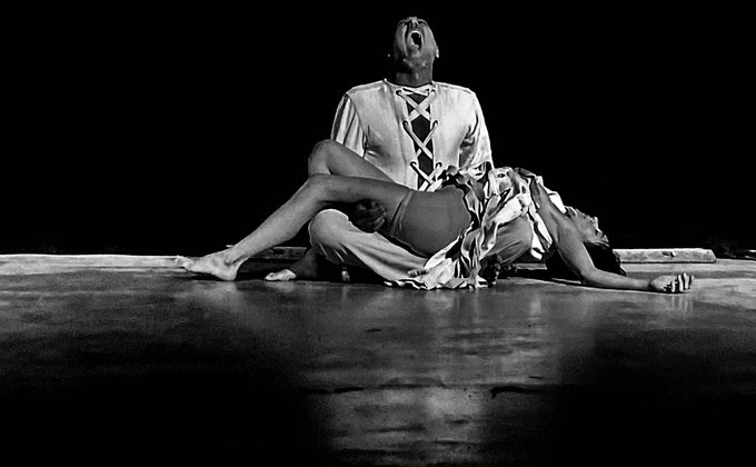 A dramatic black-and-white performance photograph captures two dancers in an emotionally charged tableau—one seated with head thrown back in an anguished cry while the other lies collapsed across their lap in a pose of surrender or grief—visually embodying the post's themes of night as lingering distance and homeland memories. The stark contrast and raw physicality reinforce the Iranian performance art piece's exploration of separation and silent endurance.