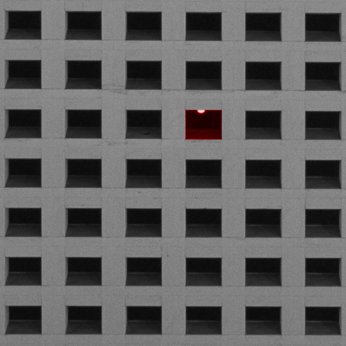 A grid-patterned concrete wall with recessed squares, where one single opening glows red with a small light, creating a stark contrast against the gray, repeating pattern.
