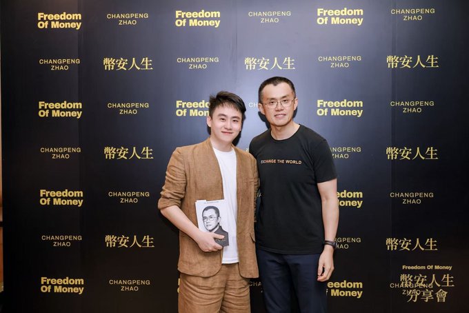 Two men pose together at a "Freedom Of Money / 幣安人生" event backdrop, with the man on the right (Changpeng Zhao, founder of Binance) standing next to a younger attendee who holds up a book featuring CZ's image. The setting matches the post's reference to CZ's Binance Life Sharing Session, where the attendee had a candid, down-to-earth interaction with the crypto industry figure.