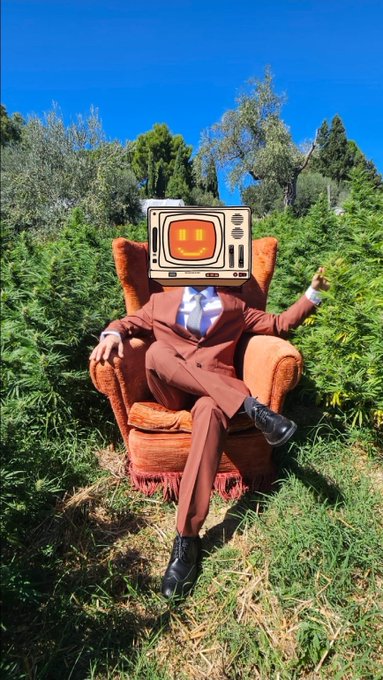 A surreal figure wearing a brown suit and tie sits in an orange armchair outdoors in a green forest, with a vintage television set displaying a smiling orange face as its head.