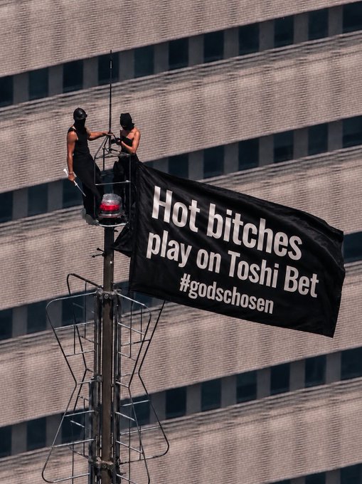 Two people stand atop an antenna structure next to a large black banner reading "Hot bitches play on Toshi Bet #godschosen," with the facade of the Empire State Building visible in the background, illustrating the stunt that led to their arrest.