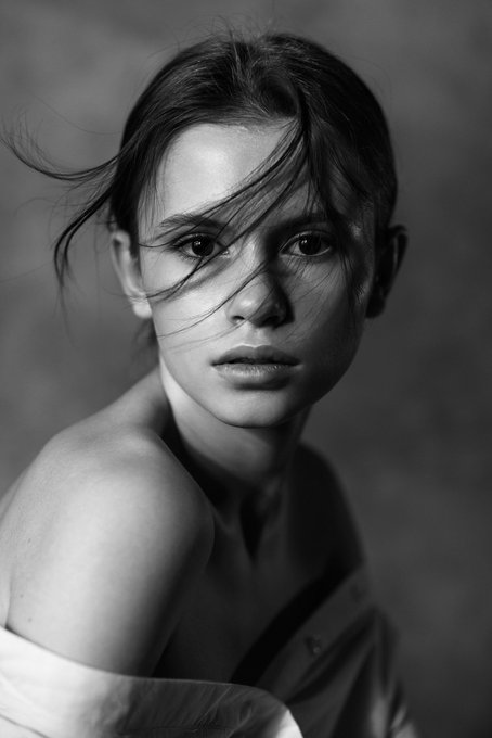 A black-and-white fashion portrait of a young woman with windswept dark hair partially covering her face, looking directly at the camera with a neutral expression.