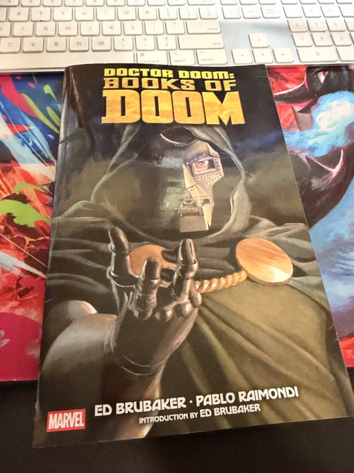 A Marvel comic book titled 'DOCTOR DOOM: BOOKS OF DOOM' by Ed Brubaker and Pablo Raimondi, featuring Doctor Doom in his hooded mask and armor on the cover, sitting on a desk next to a keyboard.