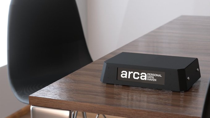 A black desktop device labeled 'arca PERSONAL DATA HAVEN' sits on a wooden table next to a black chair.
