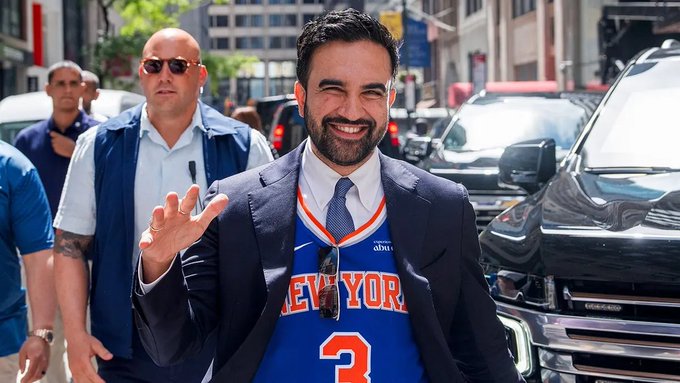 Zohran Mamdani, a candidate, smiles broadly while wearing a New York Knicks jersey under a suit jacket, walking down a city street flanked by security and an SUV.