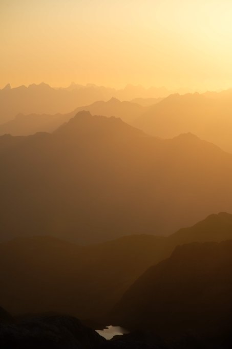 A scenic landscape photo of layered mountain silhouettes bathed in warm orange sunset or sunrise light, with a small body of water visible in the valley below.
