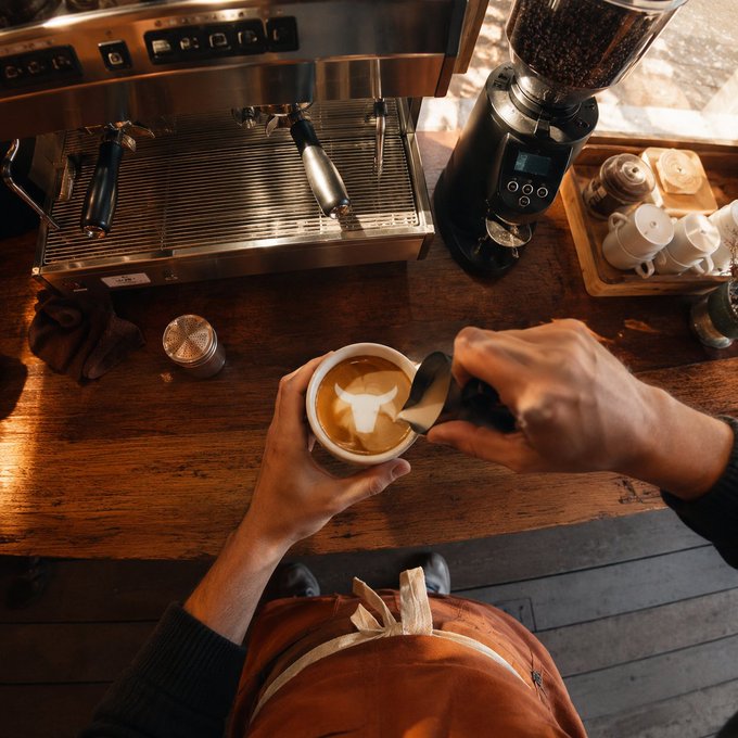 A first-person view of a barista creating latte art in a cup, with an espresso machine, coffee grinder, and a brown apron visible in the warm-lit coffee shop.